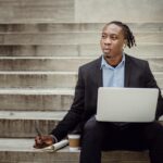 thoughtful black worker using netbook and taking notes sitting on steps