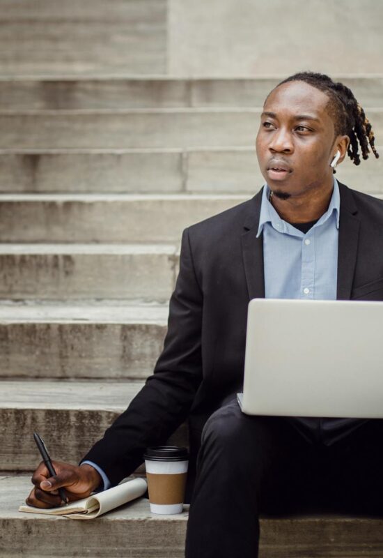 thoughtful black worker using netbook and taking notes sitting on steps