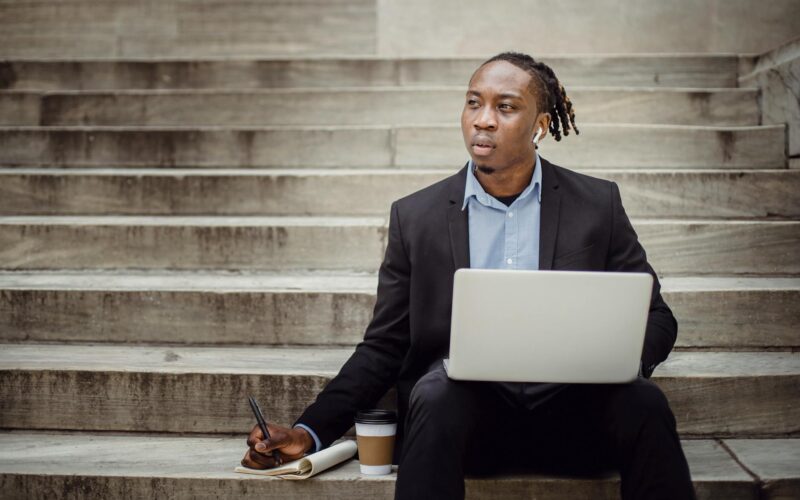 thoughtful black worker using netbook and taking notes sitting on steps